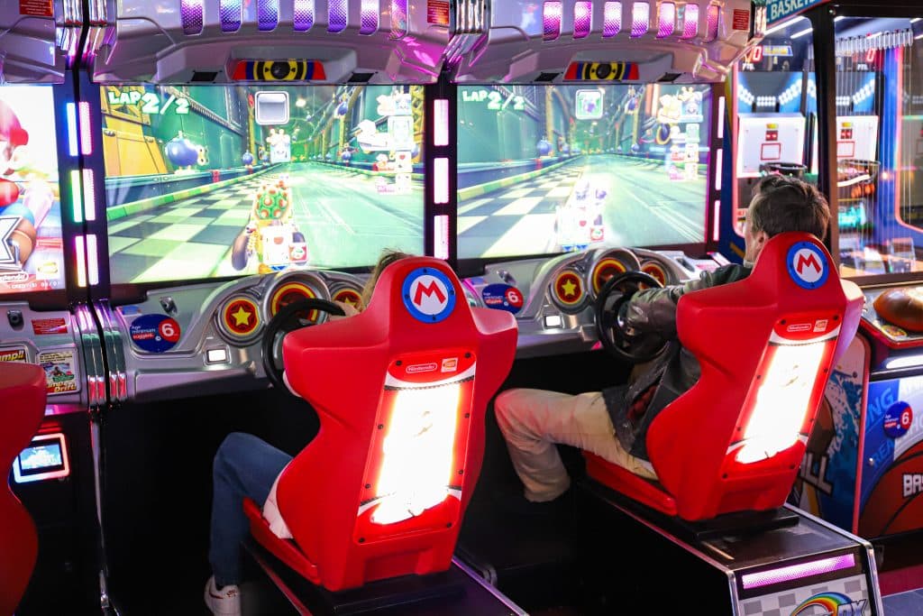 Deux personnes jouant à Mario Kart Arcade dans une salle de jeux, assises sur des sièges rouges Nintendo avec volants.